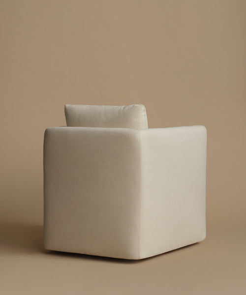 A minimalist cube-shaped upholstered armchair in a warm off-white fabric, set against a solid beige studio background.