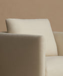 Close-up of a cream-colored upholstered armchair with a matching back cushion, set against a solid warm beige background.