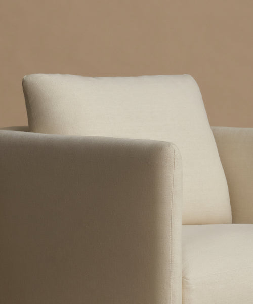 Close-up of a cream-colored upholstered armchair with a matching back cushion, set against a solid warm beige background.
