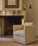 A beige upholstered armchair with a square back and thick cushions, placed in front of a stone fireplace in a warm, neutral-toned room.