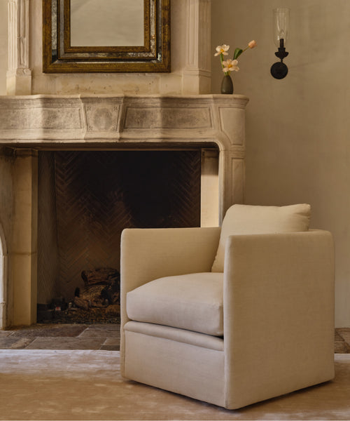 A beige upholstered armchair with a square back and thick cushions, placed in front of a stone fireplace in a warm, neutral-toned room.