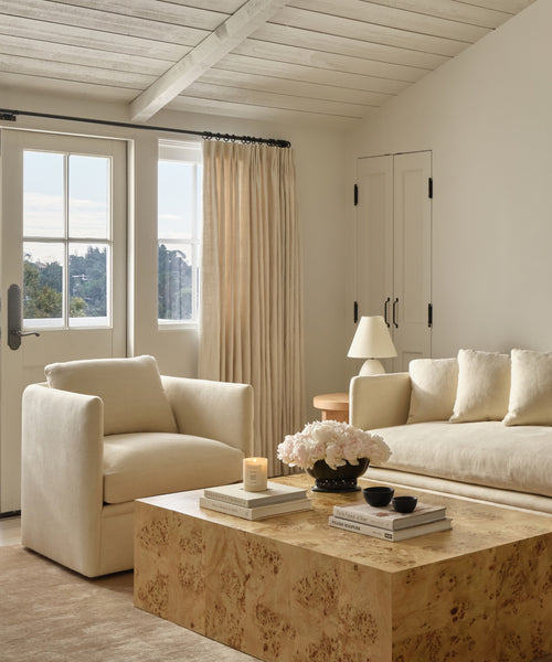 A styled living room interior featuring a cream upholstered armchair, a matching sofa, a burl wood coffee table, beige curtains, and a slatted wood ceiling.