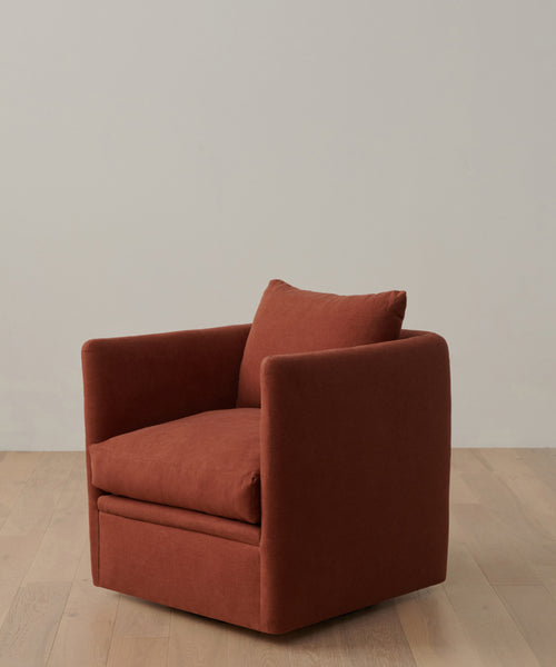 A rust-colored, modern Dume Chair with a hypoallergenic pillow sits on a light wood floor against a plain, light gray wall.