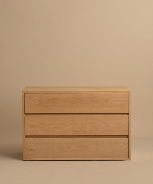 The Dune Dresser, a light White Oak piece with three wide drawers, stands against a plain beige background. Its design is minimalist and handle-free, inspired by Jenni Kayne Furniture.