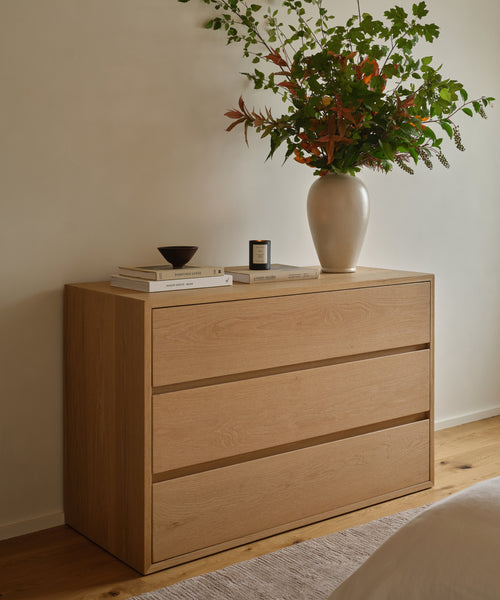 A minimal Dune Dresser in White Oak with three drawers stands against a cream wall, topped with books, a small bowl, a candle, and a vase of leafy branches for a calm, neatly arranged look.