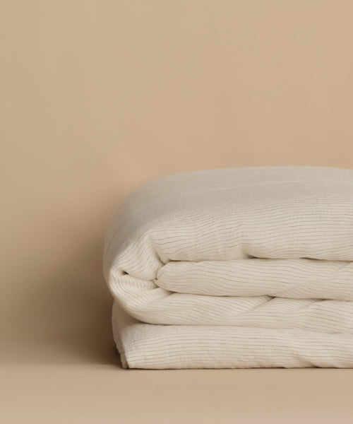 A neatly folded, cream-colored Dusk Linen Duvet Cover—crafted from lightweight, ribbed linen—is displayed on a beige surface with a matching beige background.