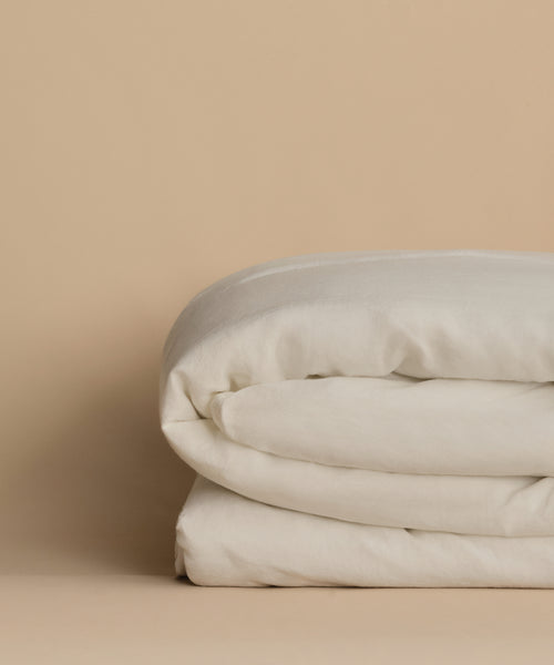 The Dusk Linen Duvet Cover, in off-white, is neatly folded on a beige surface with a matching background, highlighting its luxury and understated elegance.