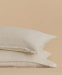 The Dusk Linen Pillow Sham Set of 2, shown in light beige with subtle flax stripes, is stacked against a tan backdrop. Made in Portugal, these shams feature soft, lightly textured fabric for a cozy look and feel.