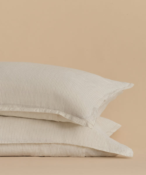 The Dusk Linen Pillow Sham Set of 2, shown in light beige with subtle flax stripes, is stacked against a tan backdrop. Made in Portugal, these shams feature soft, lightly textured fabric for a cozy look and feel.