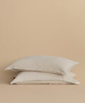 The Dusk Linen Pillow Sham Set of 2, crafted in Portugal with white linen and subtle flax stripes, is stacked neatly against a plain beige background.