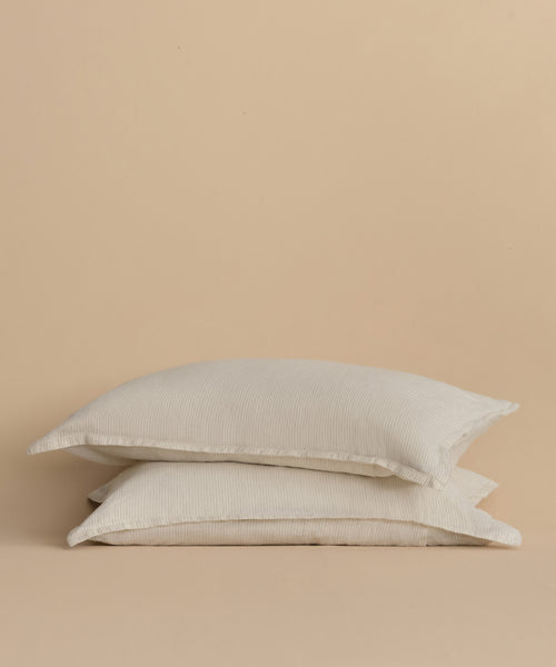 The Dusk Linen Pillow Sham Set of 2, crafted in Portugal with white linen and subtle flax stripes, is stacked neatly against a plain beige background.