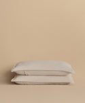 Two Dusk Linen Pillow Shams, in light beige and crafted from lightweight linen, are stacked neatly against a matching beige background.