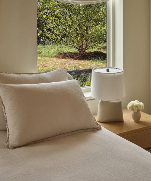 A cozy bedroom with a beige bed and Dusk Linen Pillow Sham Set of 2, a wooden nightstand holding a white ceramic lamp and small vase of white flowers, plus a window revealing green trees and grass outside.