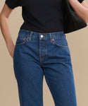A person wears a black short-sleeve shirt tucked into RE/DONE Easy Straight Crop jeans with a relaxed leg, holds a black bag under their arm, and stands against a plain beige background.