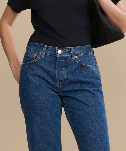 A person wears a black short-sleeve shirt tucked into RE/DONE Easy Straight Crop jeans with a relaxed leg, holds a black bag under their arm, and stands against a plain beige background.