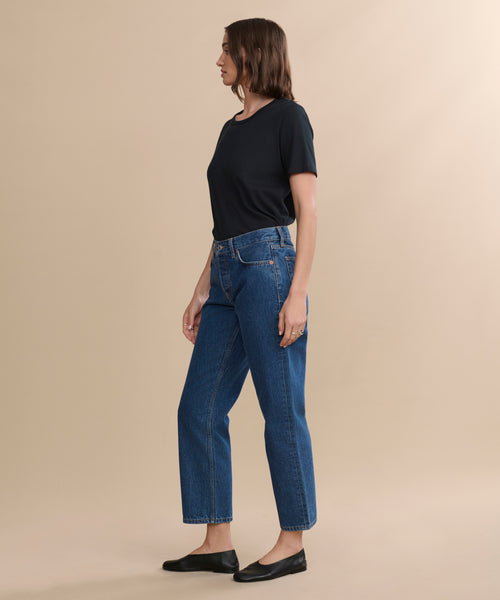 A woman in profile stands against a plain beige background, wearing a black short-sleeve shirt, blue Easy Straight Crop jeans with a relaxed leg, and black flats.