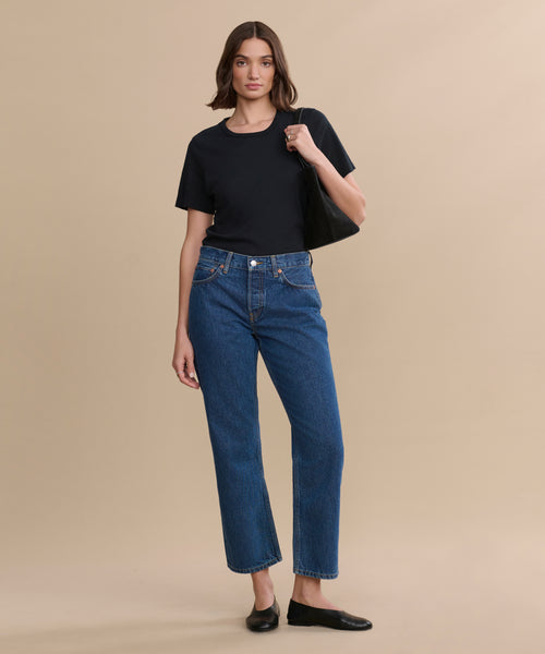 A woman stands against a beige background, wearing a black t-shirt, RE/DONE Easy Straight Crop blue high-waisted jeans with a relaxed leg, black flats, and holding a black shoulder bag.