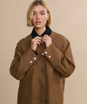 A woman with short blonde hair wears the Eaton Jacket, a garment-dyed canvas chore coat with a black collar and white buttons, standing against a beige background as she adjusts the collar and looks at the camera.