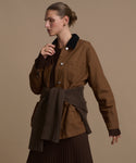 A person wearing the Eaton Jacket—a brown garment-dyed canvas chore coat with a black collar—paired with a ribbed brown sweater tied at the waist and a pleated brown skirt, stands against a plain beige background.