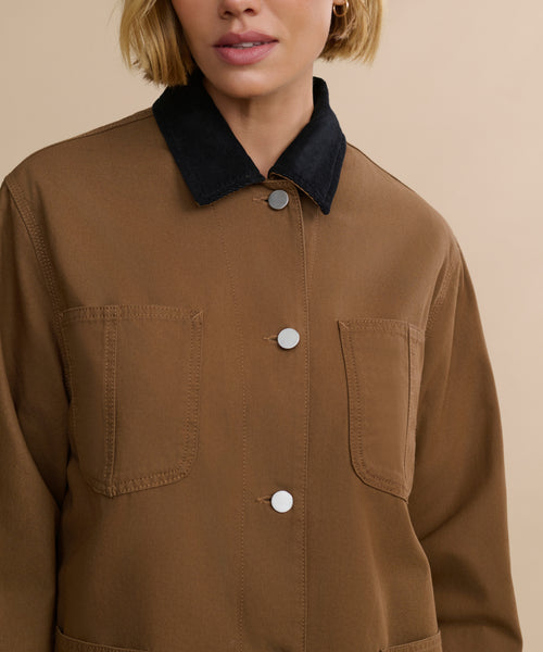 A woman models the Eaton Jacket, a brown chore coat-style jacket with a black collar, silver buttons, and two chest pockets. She stands against a beige background with her blonde bob visible and face partially shown.