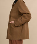 A person wearing the brown Eaton Jacket—a garment-dyed canvas chore coat with large pockets—stands sideways with one hand in a pocket, set against a beige background.