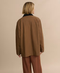 A person with shoulder-length blonde hair, wearing the Eaton Jacket and matching pants in brown, stands with their back to the camera against a plain beige background.
