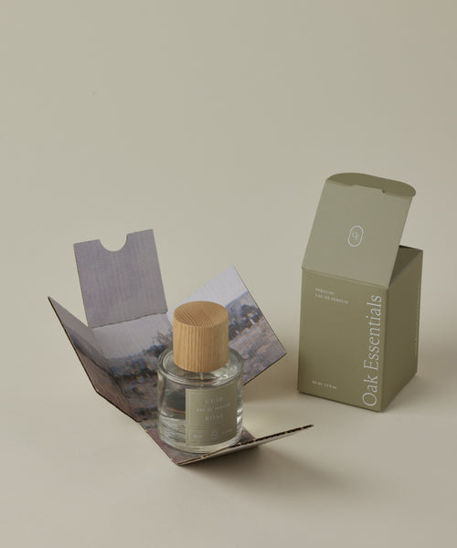 A glass bottle of Cuir Rose Eau De Parfum with a wooden cap rests in an open box with scenic artwork, beside a closed olive-green Oak Essentials box on a beige background, evoking the floral and woody notes of this clean fragrance.