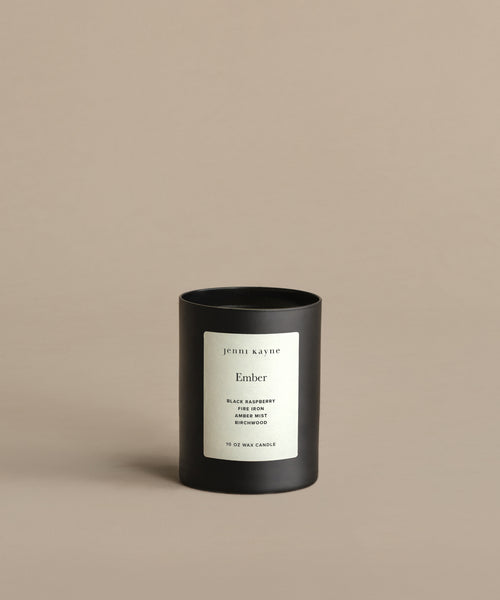 A black Ember Glass Candle with a white “jenni kayne Ember” label rests on a beige background. Hand-poured in Los Angeles, this natural candle features scent notes of black raspberry, plum, patchouli, birchwood, and birch.