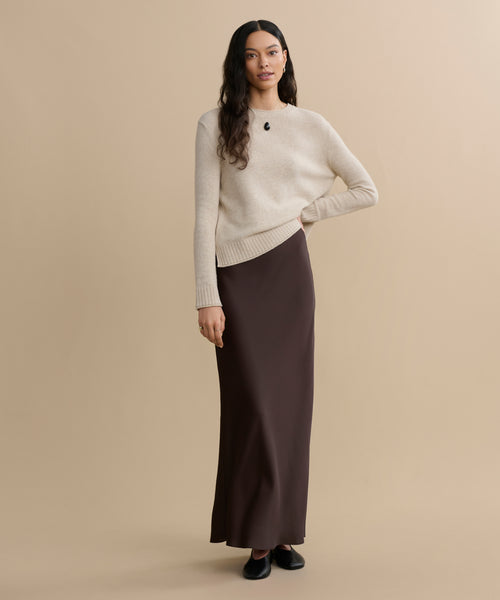 A woman with long wavy hair stands against a beige background, wearing the Everyday Sweater in cream, paired with a long brown skirt and black shoes. Her arms are relaxed at her sides and she has a calm expression.