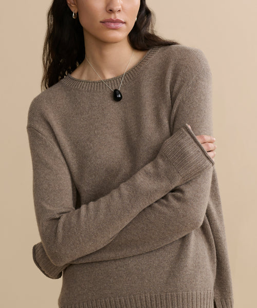 A person with long dark hair stands with arms crossed against a beige background, wearing the Everyday Sweater and a black pendant necklace.