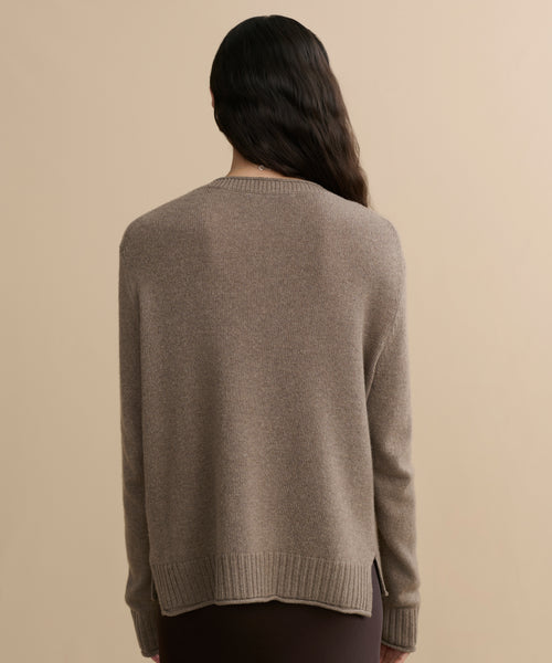 A person with long, wavy dark hair is seen from behind wearing the Everyday Sweater, a loose-fitting taupe knit with ribbed cuffs and hem, against a plain beige background.