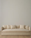 A modern sofa upholstered in Fabric Swatch Beige Hemp, accented with five matching cushions, sits against a plain light wall on a wooden floor.