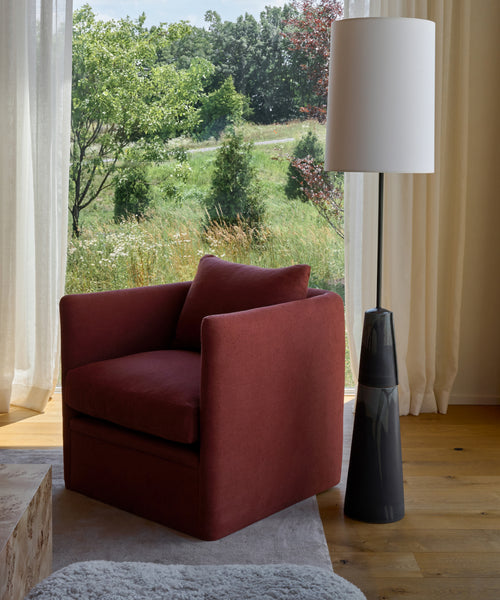 A modern armchair in Farmhouse Red Linen sits beside a tall black floor lamp by a large window with sheer curtains, offering views of a sunlit landscape with trees and grass.