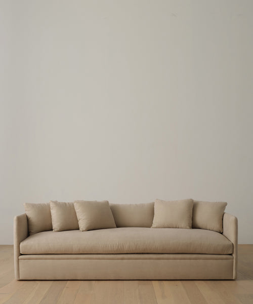 A beige Dume Sofa in Sand Hemp fabric, paired with four matching cushions, sits on a light wooden floor by an off-white wall. The look is completed with the Fabric Swatch Sand Hemp for a refined modern touch.