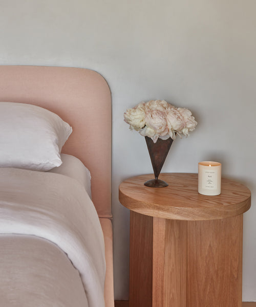 A Cove Bed dressed in Fabric Swatch Shell Linen stands near a wooden nightstand with a white candle and a metal vase of white and pale pink flowers, set against a light gray wall.