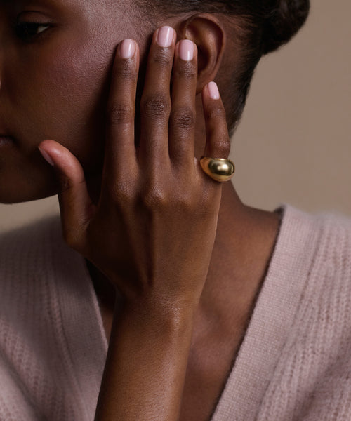 A person with dark skin, wearing a soft light pink sweater, shows off their manicured nails and the Felix Dome Ring against a neutral background.