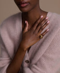 A woman in a pale pink cardigan gently places her hand on her chest, her manicured nails and the Felix Dome Ring gleaming softly against a neutral background.
