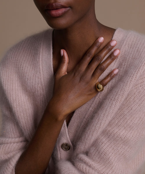 A woman in a pale pink cardigan gently places her hand on her chest, her manicured nails and the Felix Dome Ring gleaming softly against a neutral background.