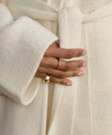 A close-up of a hand wearing the Felix Dome Ring in 14k solid gold and a thin silver band, resting on the waist of a person dressed in a soft, cream-colored belted coat from Jenni Kayne Jewelry.