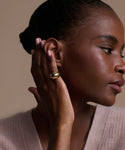 A woman with smooth brown skin poses in profile, wearing the Felix Dome Ring on her hand. She pairs the chunky gold ring with a soft pink knit sweater; her pale manicure and neatly pulled-back hair complete the look.