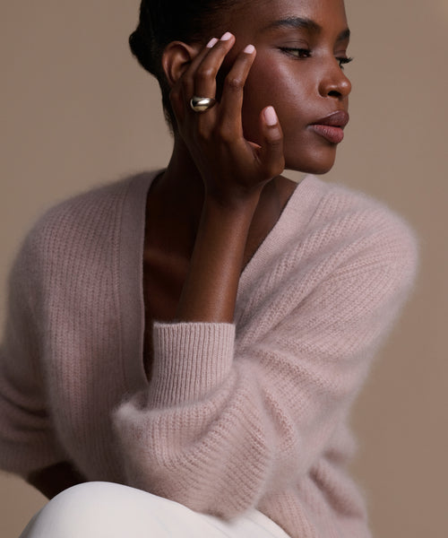 A woman wearing the Felix Dome Ring, a white gold piece, rests her chin on her hand. Dressed in a light pink sweater and white pants with manicured nails and natural makeup, she is set against a neutral background, gazing thoughtfully to the side.
