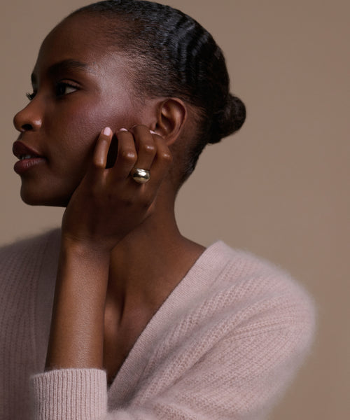 A woman with smooth dark skin and a low bun looks to the side, resting her chin on her hand to showcase the Felix Dome Ring in 14k solid gold. She wears a soft light pink sweater against a beige background.