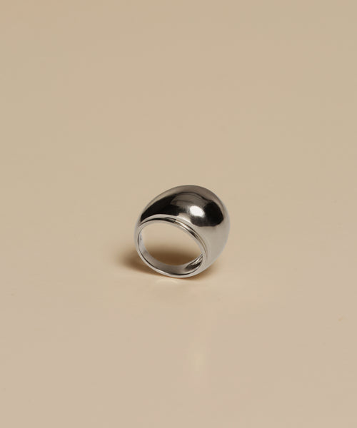 The Felix Dome Ring, a shiny silver dome-shaped white gold piece, rests on a plain beige surface and reflects light with its smooth, polished finish.