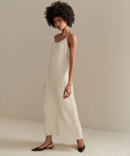 A woman with natural curly hair and dark pointed-toe flats stands on a light floor, wearing the Fete Slip Dress—an ankle-length, sleeveless, light beige dress with thin straps. She looks at the camera against a plain, neutral background.
