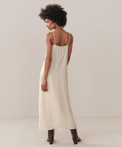A woman with short curly hair models the Fete Slip Dress—an ankle-length, sleeveless cream dress with thin straps. She stands facing away from the camera in black heeled boots against a neutral background.