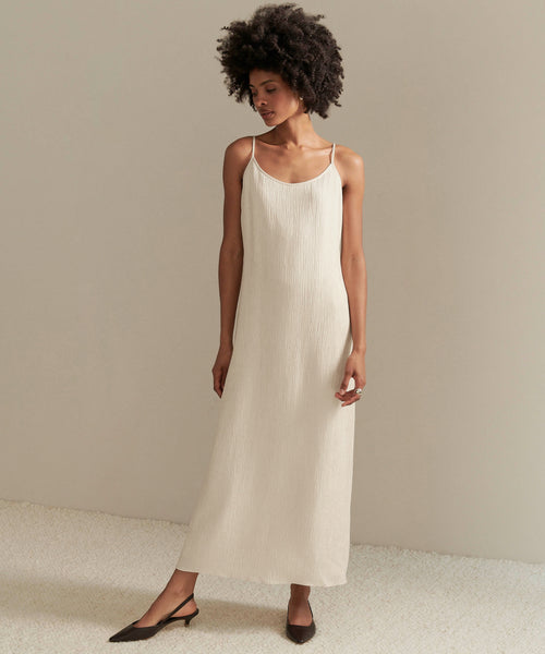 A woman with natural curly hair wears the sleeveless, ankle-length off-white Fete Slip Dress with a subtle crinkle effect and black pointed-toe low heels, standing against a plain light background.