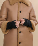 A person shown from the shoulders down wears a tan shearling coat with large buttons, several gold rings, and black Fingerless Cashmere Gloves.
