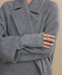 A person in a cozy gray wool coat and Fingerless Cashmere Gloves crosses their arms, displaying manicured nails and gold rings against a neutral beige background.