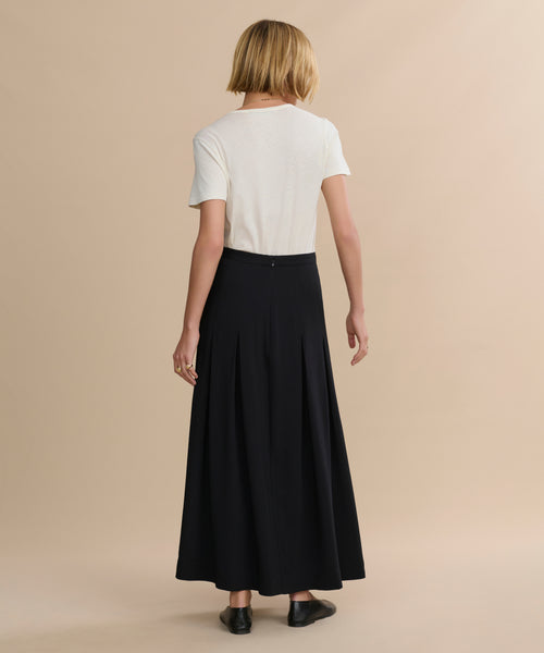 A person with short blonde hair faces away, wearing a white T-shirt, the Florence Skirt, and black shoes against a plain beige background.