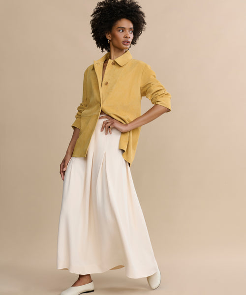 A woman poses against a beige backdrop, wearing the Florence Skirt—an elegant, flowing cream midi skirt—paired with a mustard-yellow jacket and white shoes, one hand on her hip as she gazes to the side.